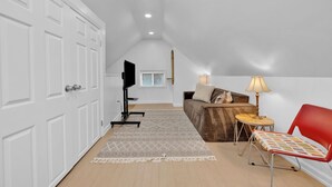Ein Zimmer mit weißer Tür, einem Fernseher auf einem Schrank, einer Couch mit Kissen, einem kleinen Tisch mit Lampe und einem roten Sessel.