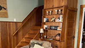 Una sala de estar acogedora con una escalera de madera, un sillón beige, un tapete estampado y una estantería llena de libros y objetos decorativos.