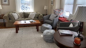 Una sala de estar con sofá, sillón, reposapiés, mesa de centro y una ventana con cortinas.