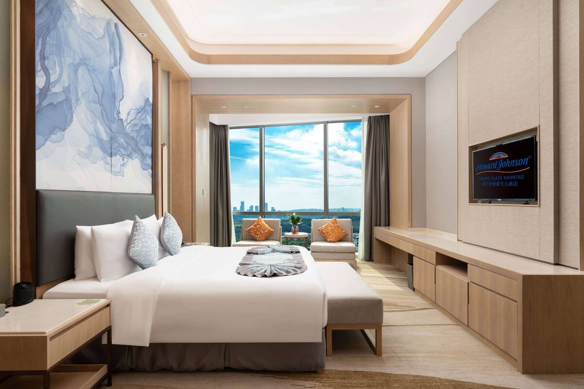 Una habitación de hotel moderna con una cama grande, televisor, vista a la ciudad y un cuadro abstracto de gran tamaño.
