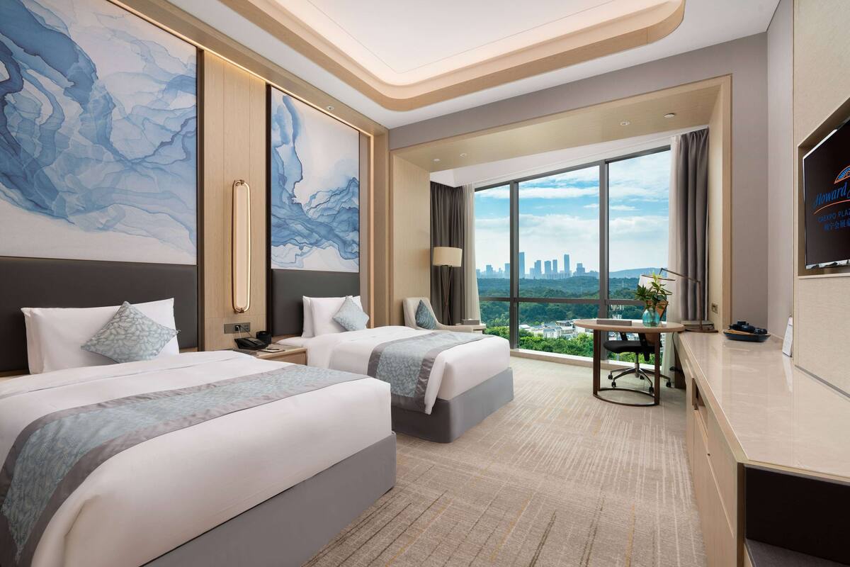 Habitación de hotel con dos camas, un escritorio, una silla, un televisor y un amplio ventanal con vistas a la ciudad.