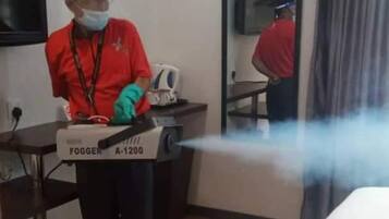 A hotel room with a bed, a desk, and a mirror. A person is operating a fogger device.
