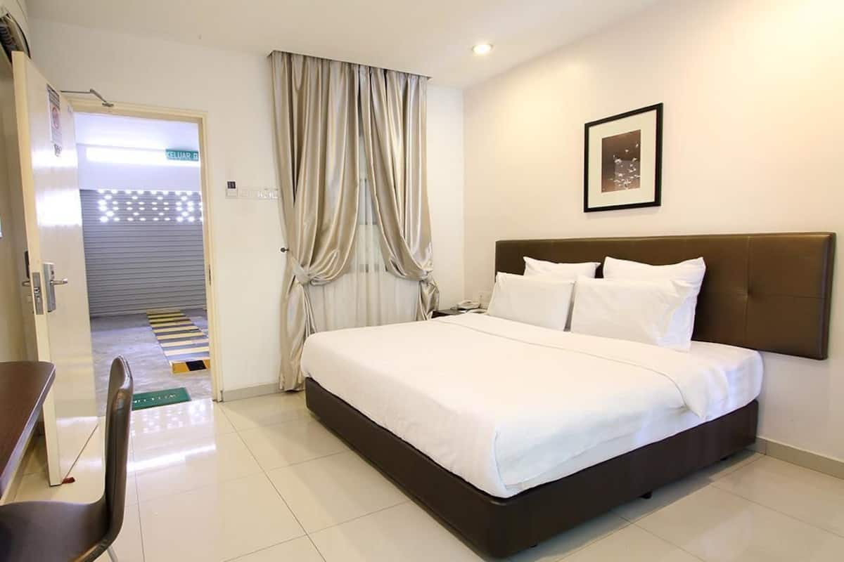 A hotel room with a bed, a chair, a desk, a framed picture on the wall, and a door leading to a corridor.