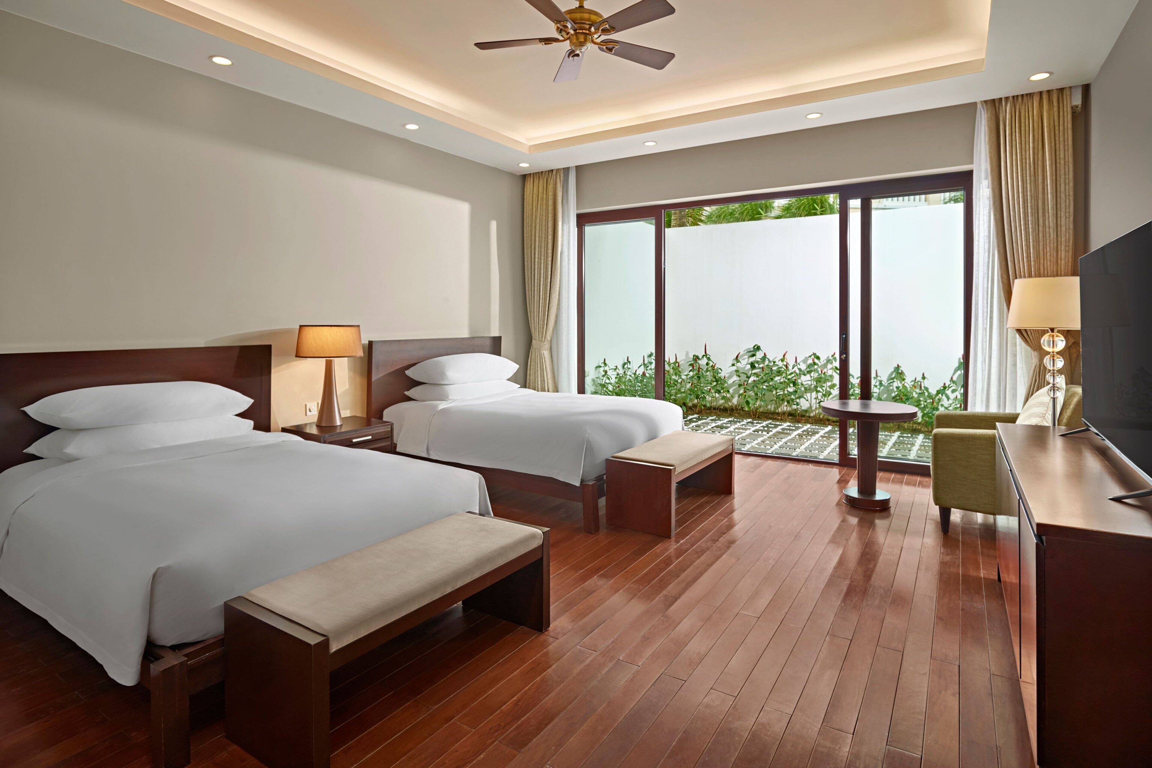 A hotel room with two beds, a desk, a chair, a TV, and a balcony with a view of greenery.