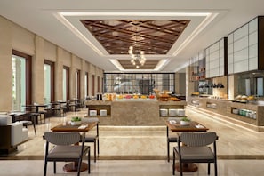 A spacious hotel lobby with a central seating area, a modern kitchen with a buffet, and large windows allowing natural light.