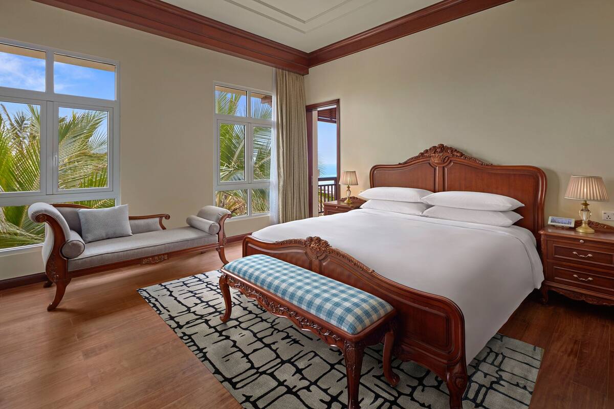 A spacious bedroom with a large bed, a bench, a sofa, and a view of palm trees through the windows.