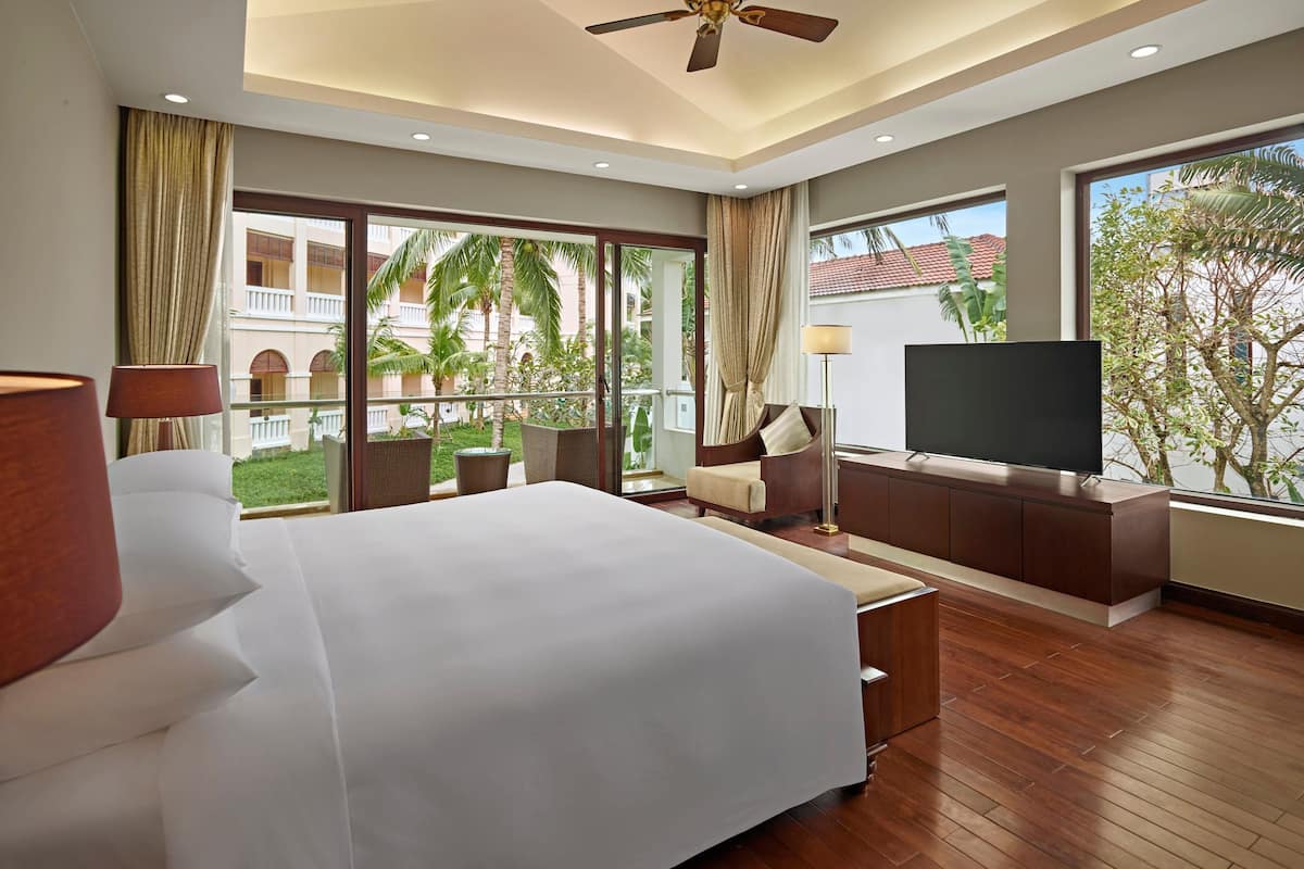 A modern hotel room with a large bed, a flat-screen TV, a balcony with a view of palm trees, and a ceiling fan.
