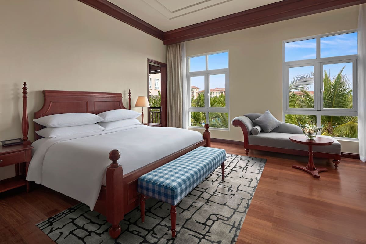 A hotel room with a large bed, a bench, a sofa, a table, and large windows with a view of palm trees.