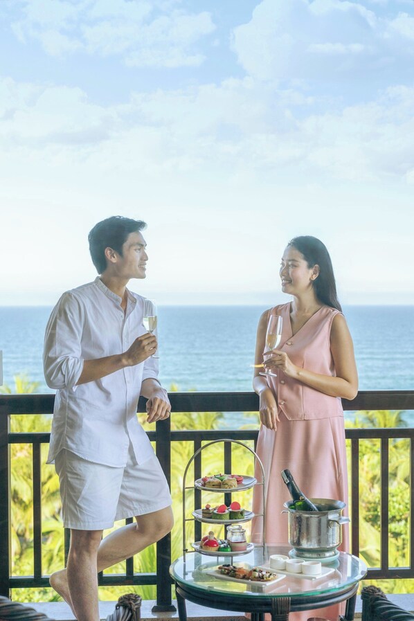 A couple standing on a balcony with a view of the sea, holding drinks, and a table with food and a bottle of champagne.