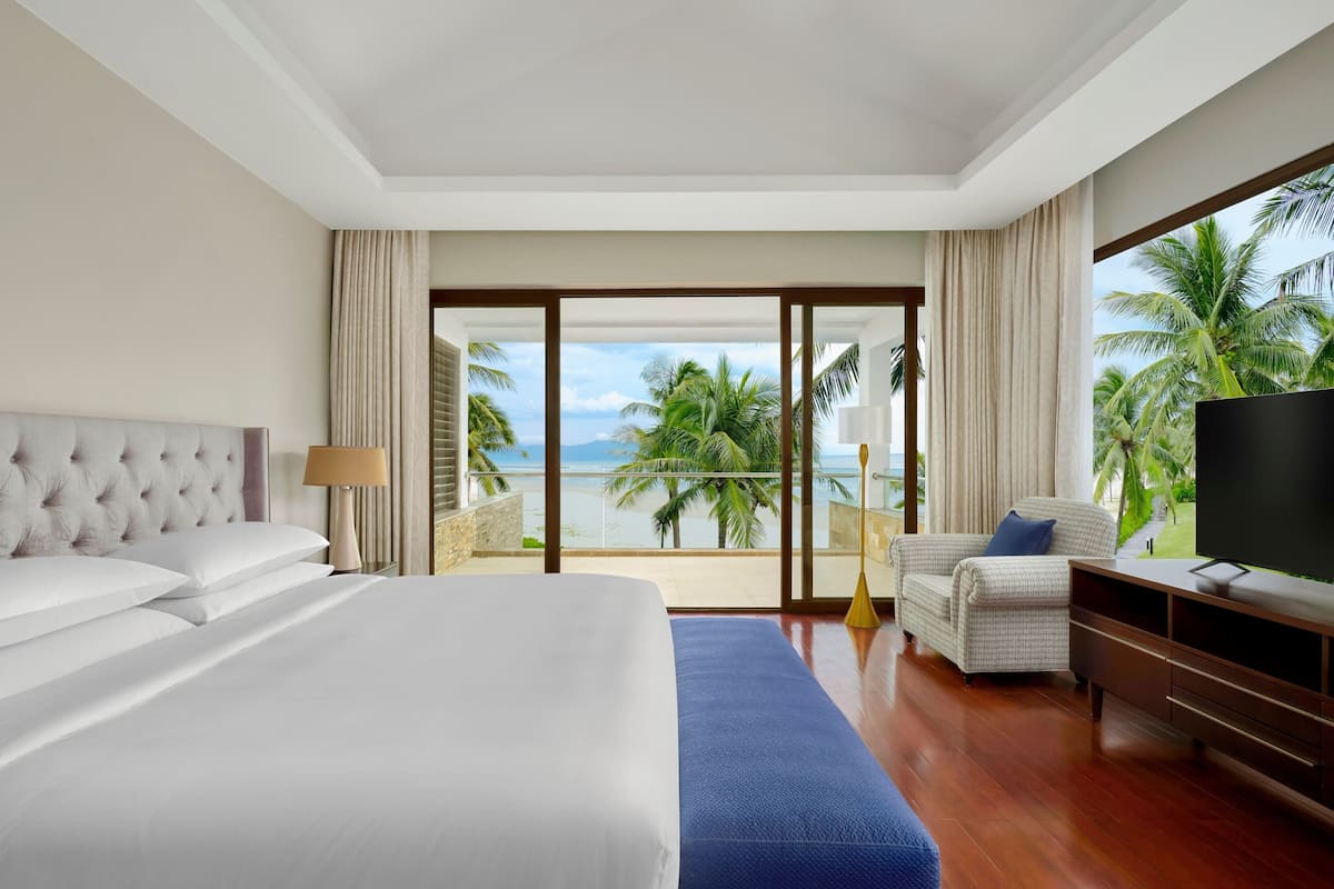 A hotel room with a large bed, a TV, a sofa, and a balcony with a view of palm trees and the ocean.