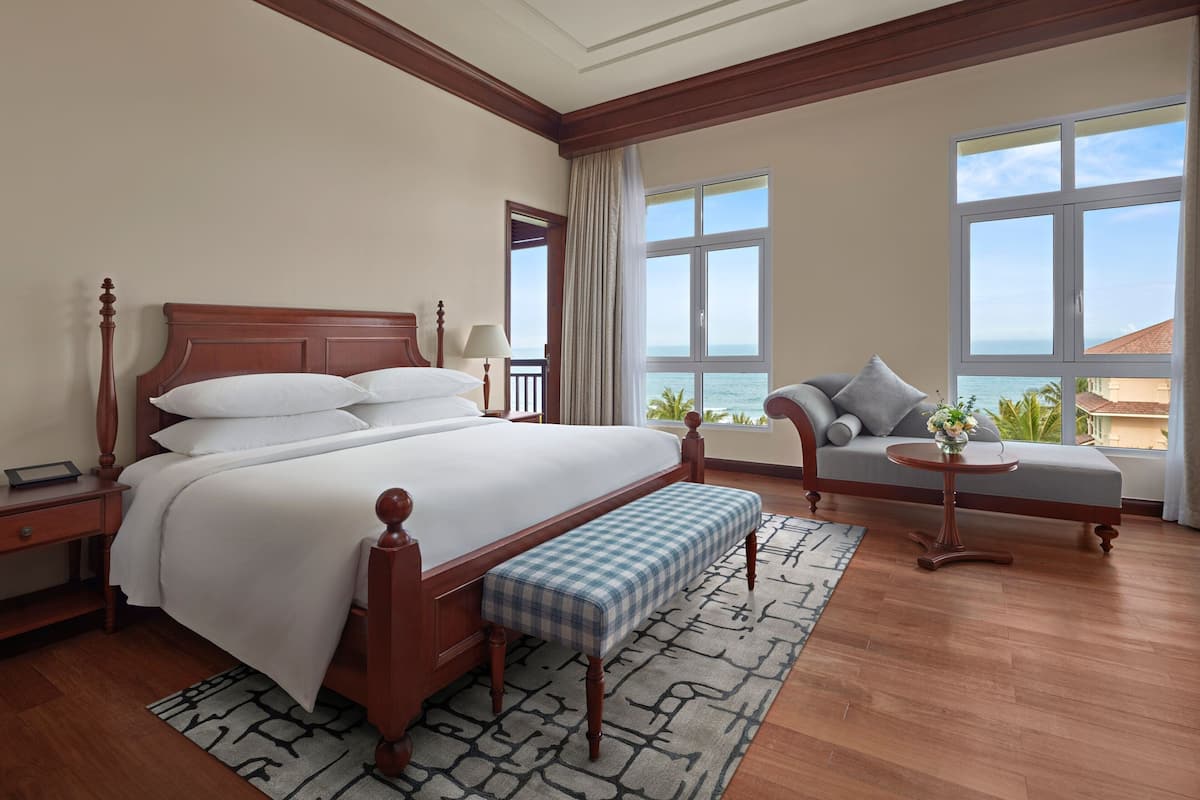 A hotel room with a large bed, a bench, a sofa, a small table, and a view of the ocean.
