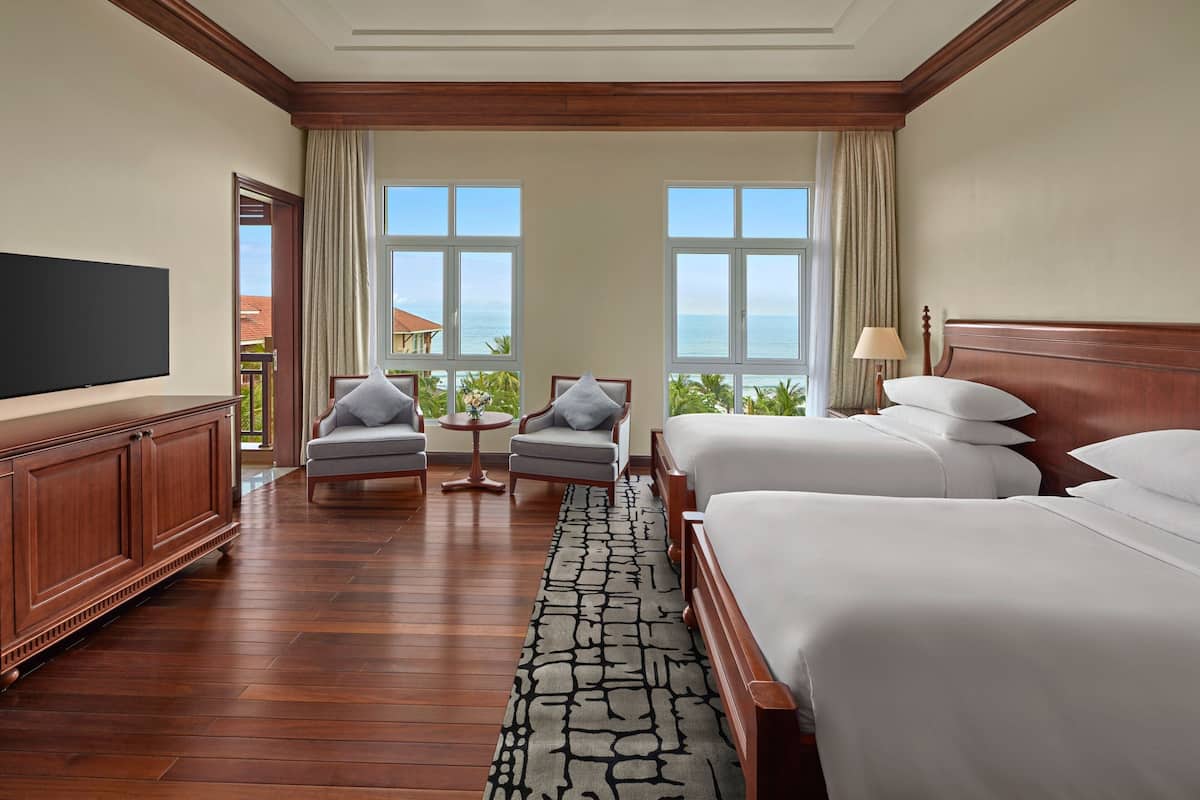 A hotel room with two beds, a TV, a wooden cabinet, a small table, and a view of the ocean.