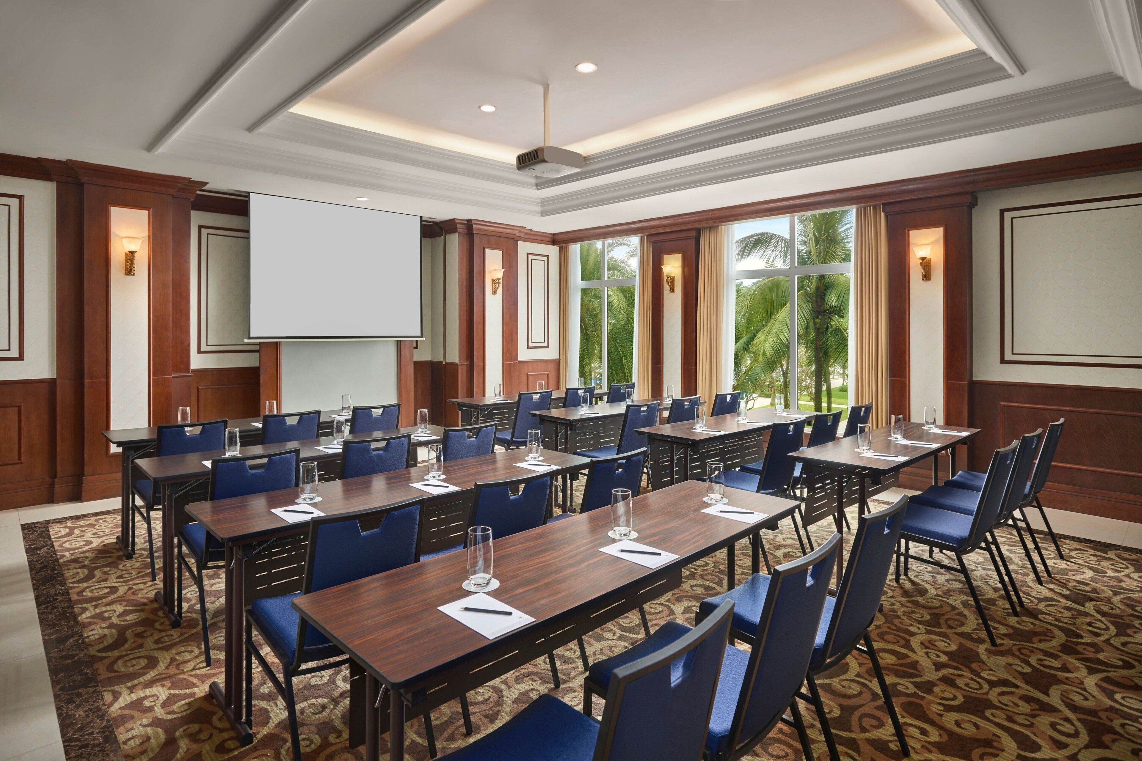 A conference room with multiple tables and chairs, a projector screen, and large windows with a view of palm trees.