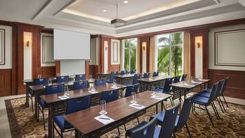A conference room with multiple tables and chairs, a projector screen, and large windows with a view of palm trees.