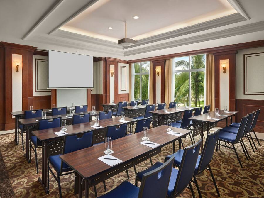 A conference room with multiple tables and chairs, a projector screen, and large windows with a view of palm trees.