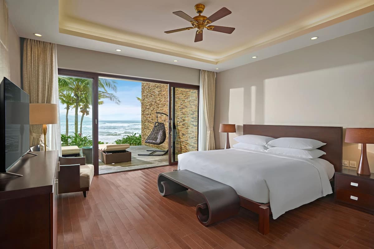 A modern hotel room with a large bed, a TV, a desk, and a view of the ocean.