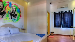 A bedroom with a bed, a bedside table, a wall with a colorful painting, a window with curtains, and a built-in shelf unit. - The Funky Village (Siem Reap)
