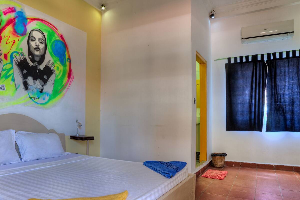 A bedroom with a bed, a bedside table, a wall with a colorful painting, a window with curtains, and a built-in shelf unit.