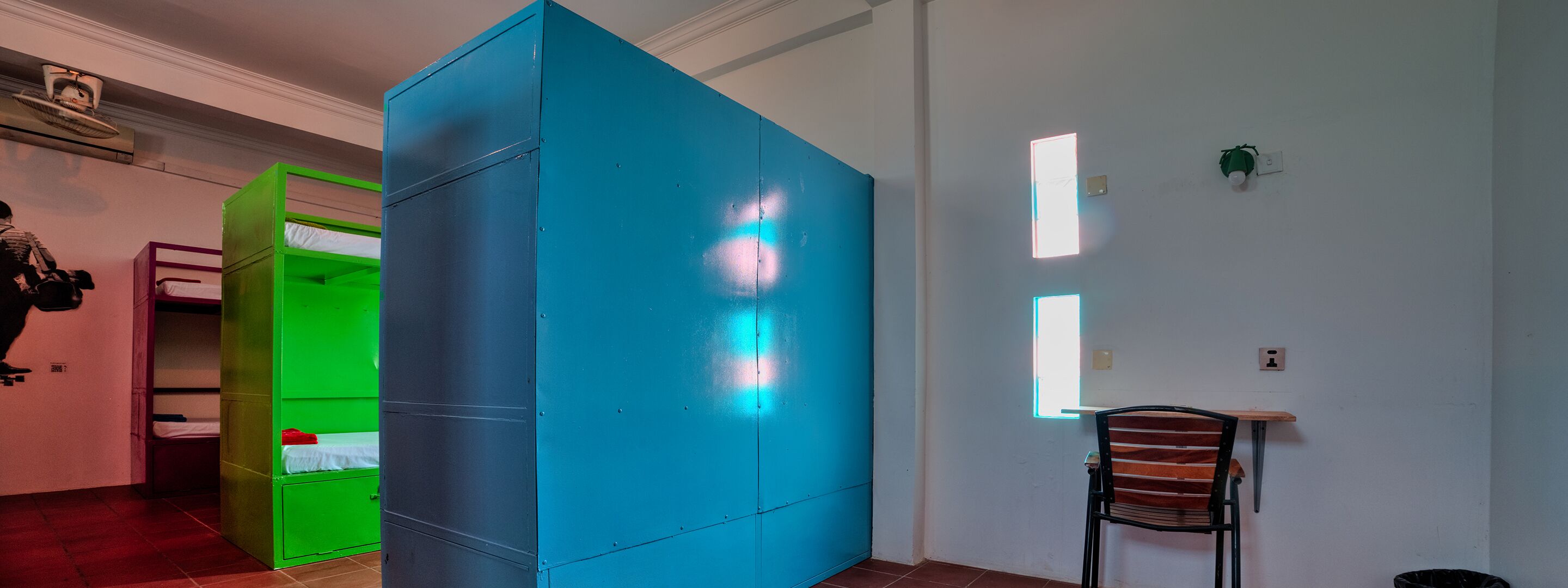 A room with a bunk bed, a green storage unit, a blue partition, and a chair.