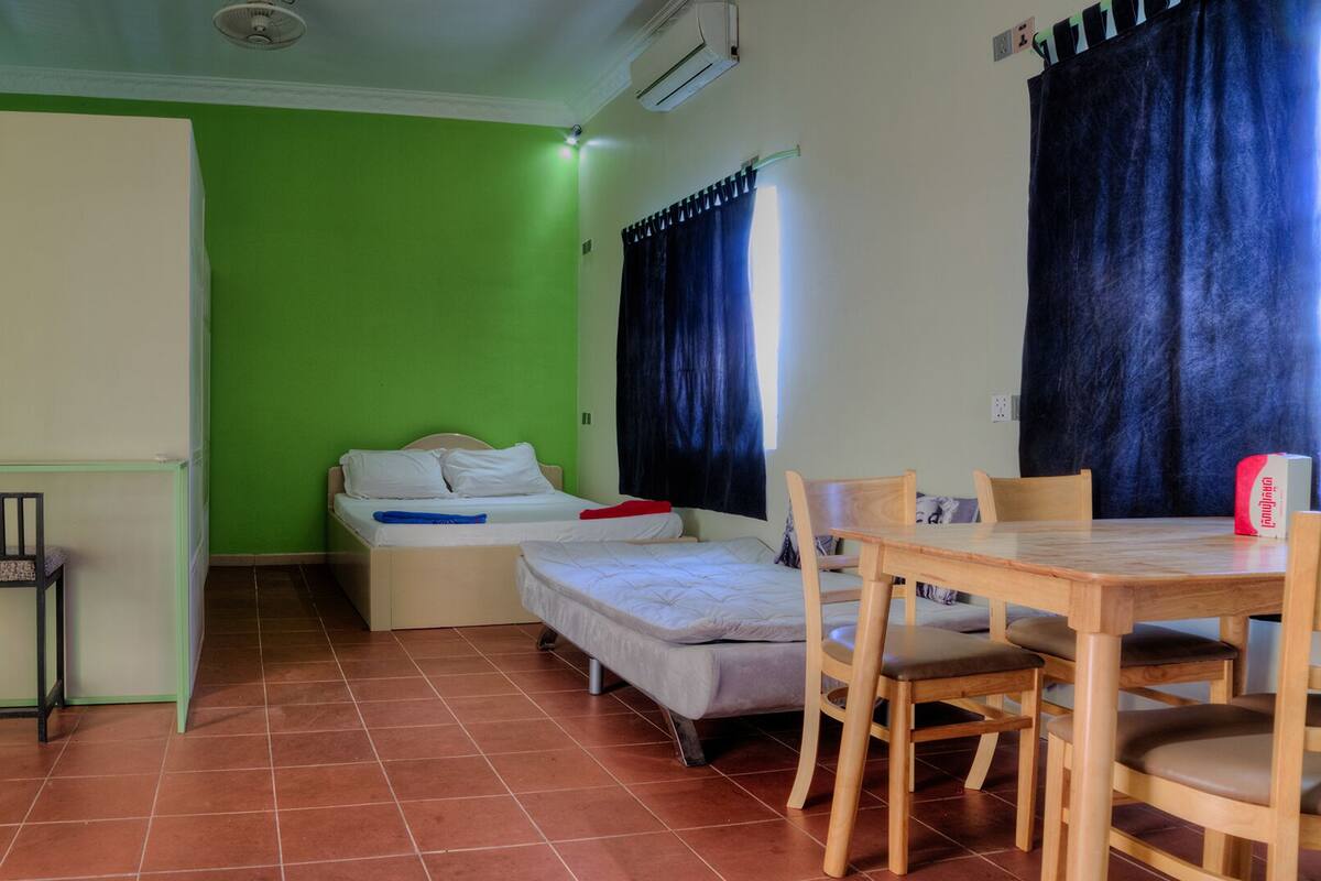 A small room with a green wall, a bed, a desk with chairs, and a small table.