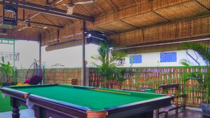 A pool table in a covered outdoor area with artificial grass, surrounded by tables and chairs, and various potted plants. - The Funky Village (Siem Reap)