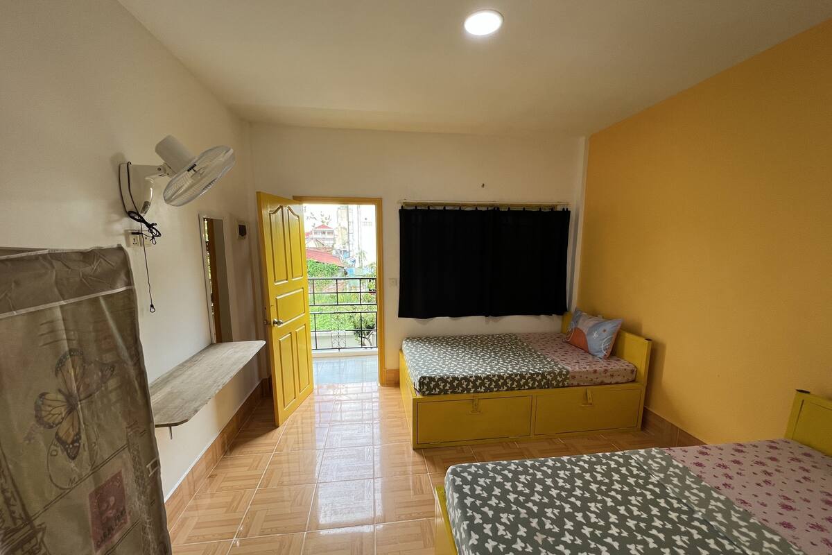 A bedroom with a bed, a bedside table, a fan, and a window with a curtain.