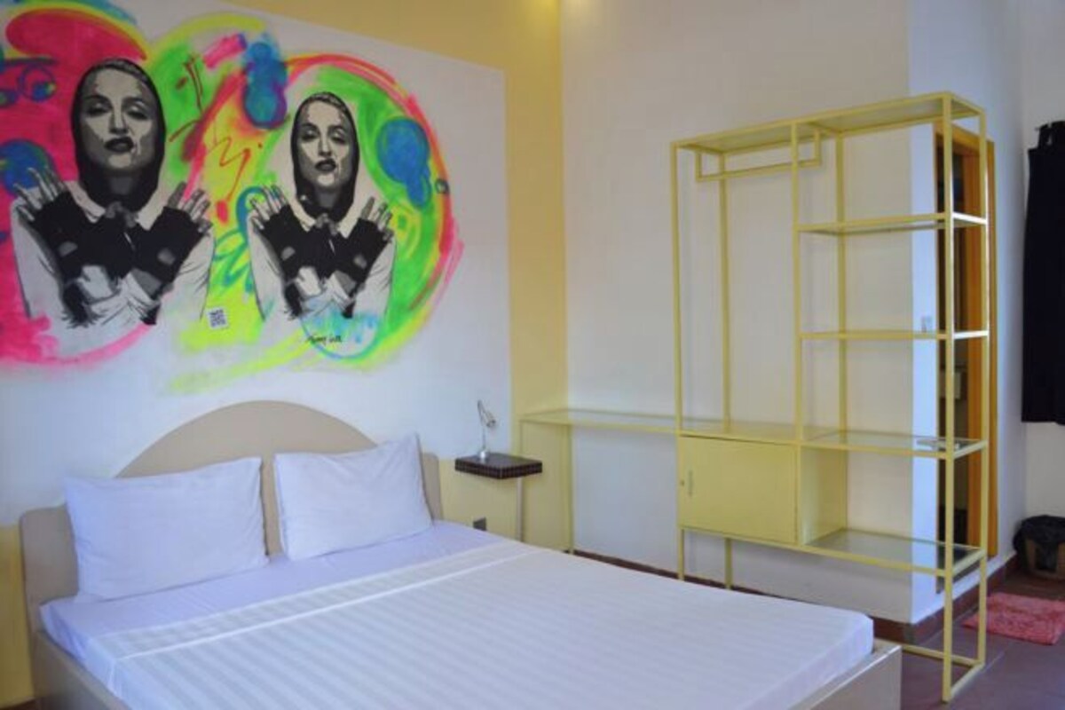 A hotel room with a bed, a white headboard, a bedside table with a lamp, and a colorful abstract painting on the wall.