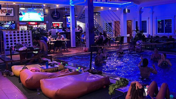 A pool area with people relaxing and a bar area with TVs and seating. - The Funky Village (Siem Reap)