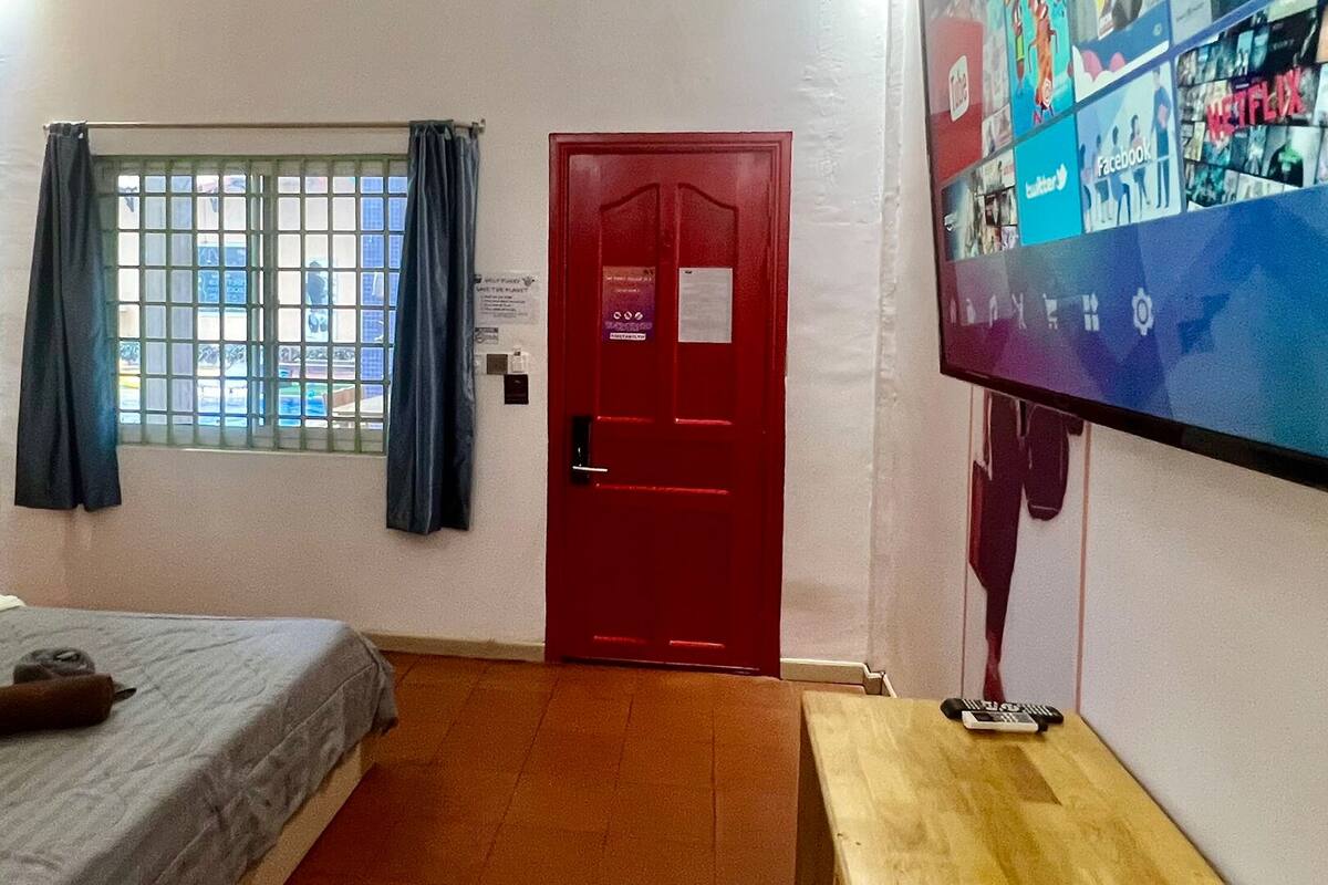 A hotel room with a bed, a TV mounted on the wall, a red door, and a window with bars.