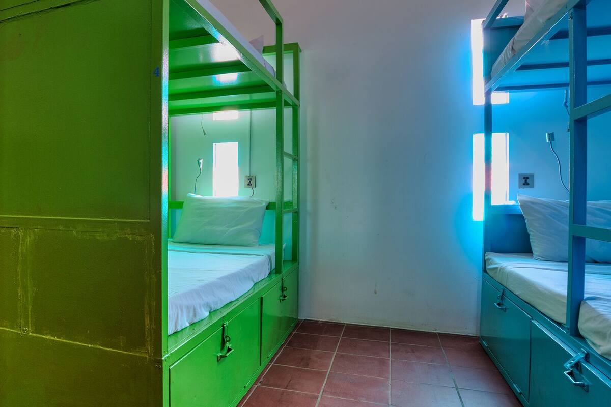 A dormitory room with bunk beds in red, green, and blue, a green wardrobe, and a window with natural light.