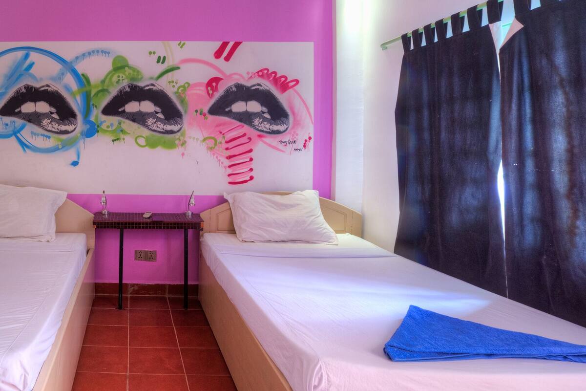 A room with two beds, a small table, a wall with graffiti art, and a bathroom with a green sink.
