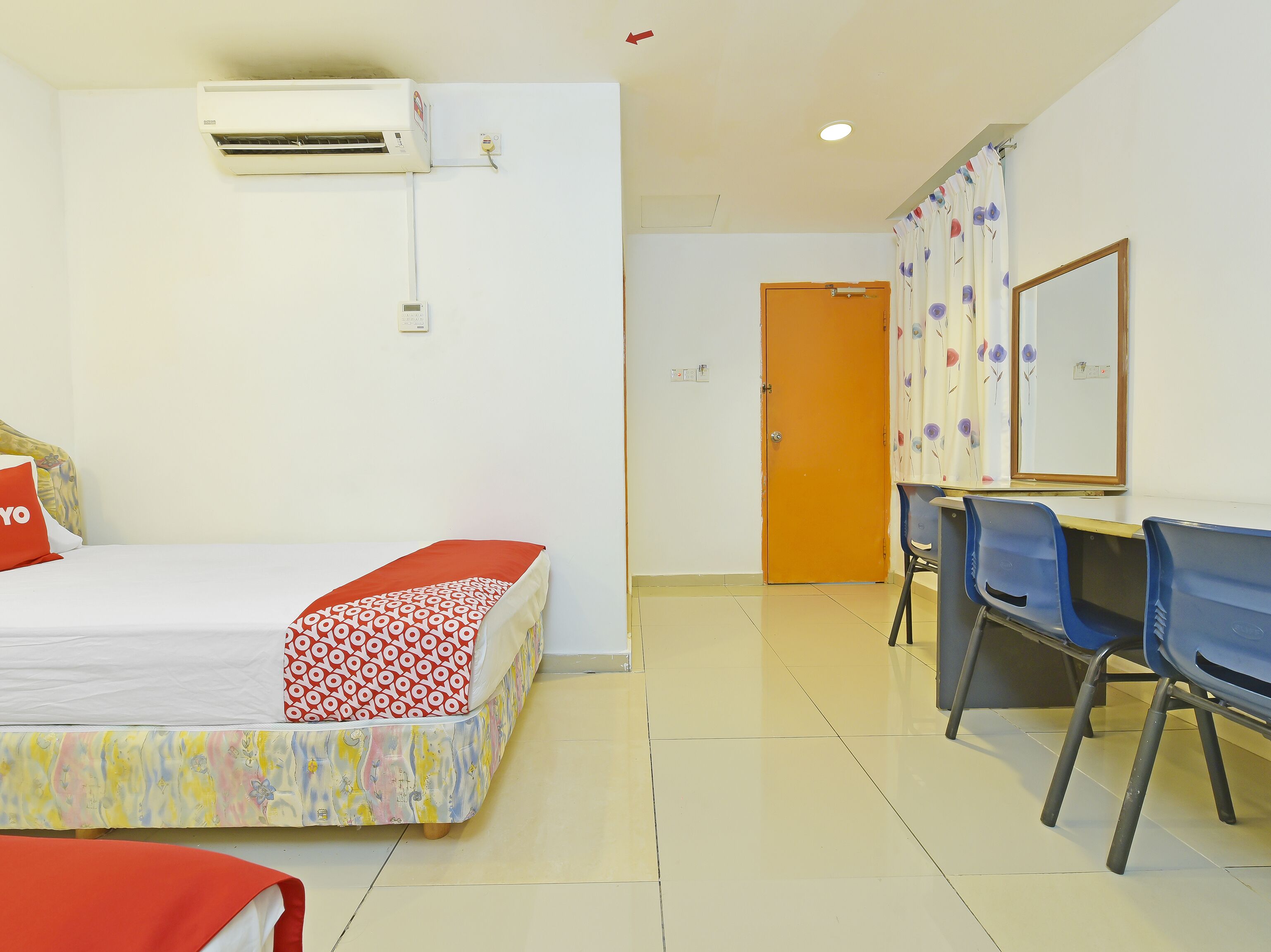 A hotel room with a bed, an air conditioning unit, a desk with chairs, and a door.