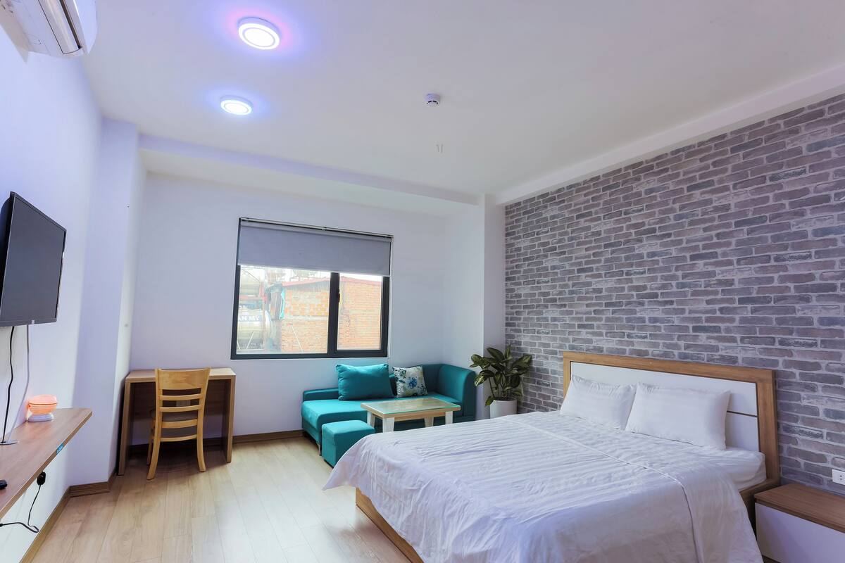 A hotel room with a bed, a desk, a chair, a small table, a sofa, and a brick-patterned wall.