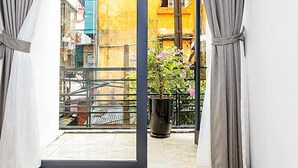 Une vue à travers une porte-fenêtre donnant sur un balcon, avec une rambarde en métal, une plante en pot et un bâtiment jaune en arrière-plan.