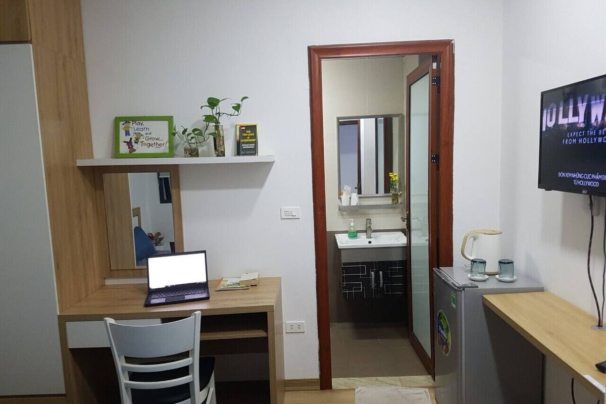 A compact hotel room with a wooden desk, a laptop, a white chair, a small refrigerator, a TV, and a bathroom visible through an open door.