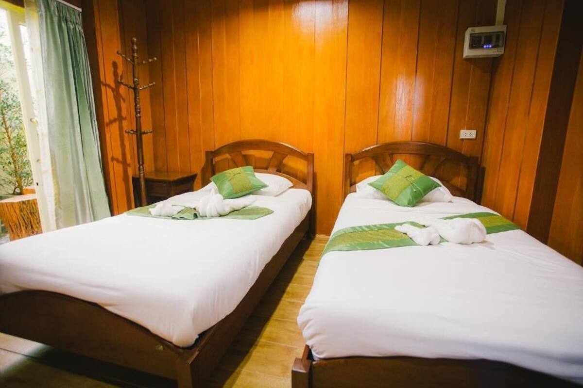 Two wooden beds with white linens and green pillows, a wooden coat rack, and a thermostat on the wall.