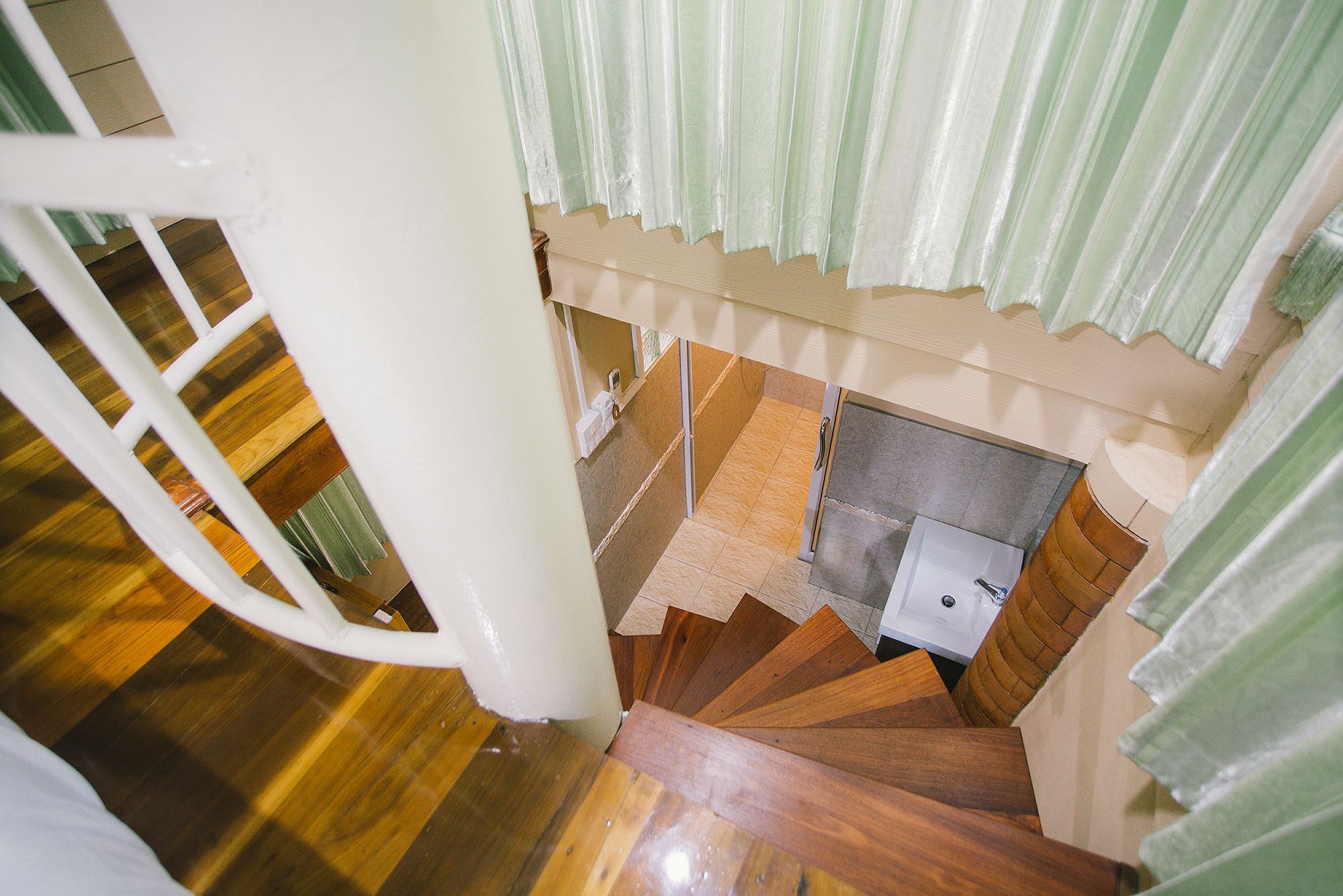 A wooden staircase with a metal railing, leading to a room with a tiled floor and a small white appliance, possibly a refrigerator.