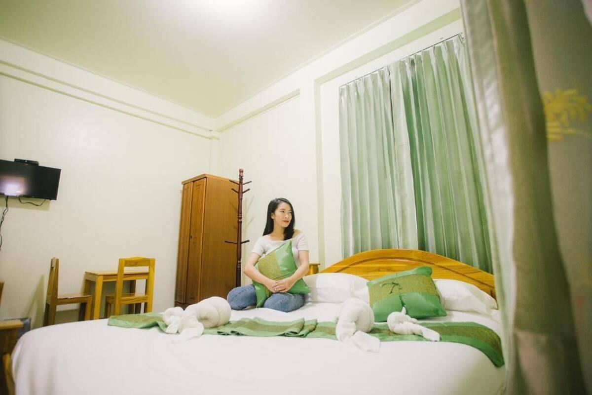 A room with a bed, a wooden wardrobe, a television mounted on the wall, and a window with curtains.