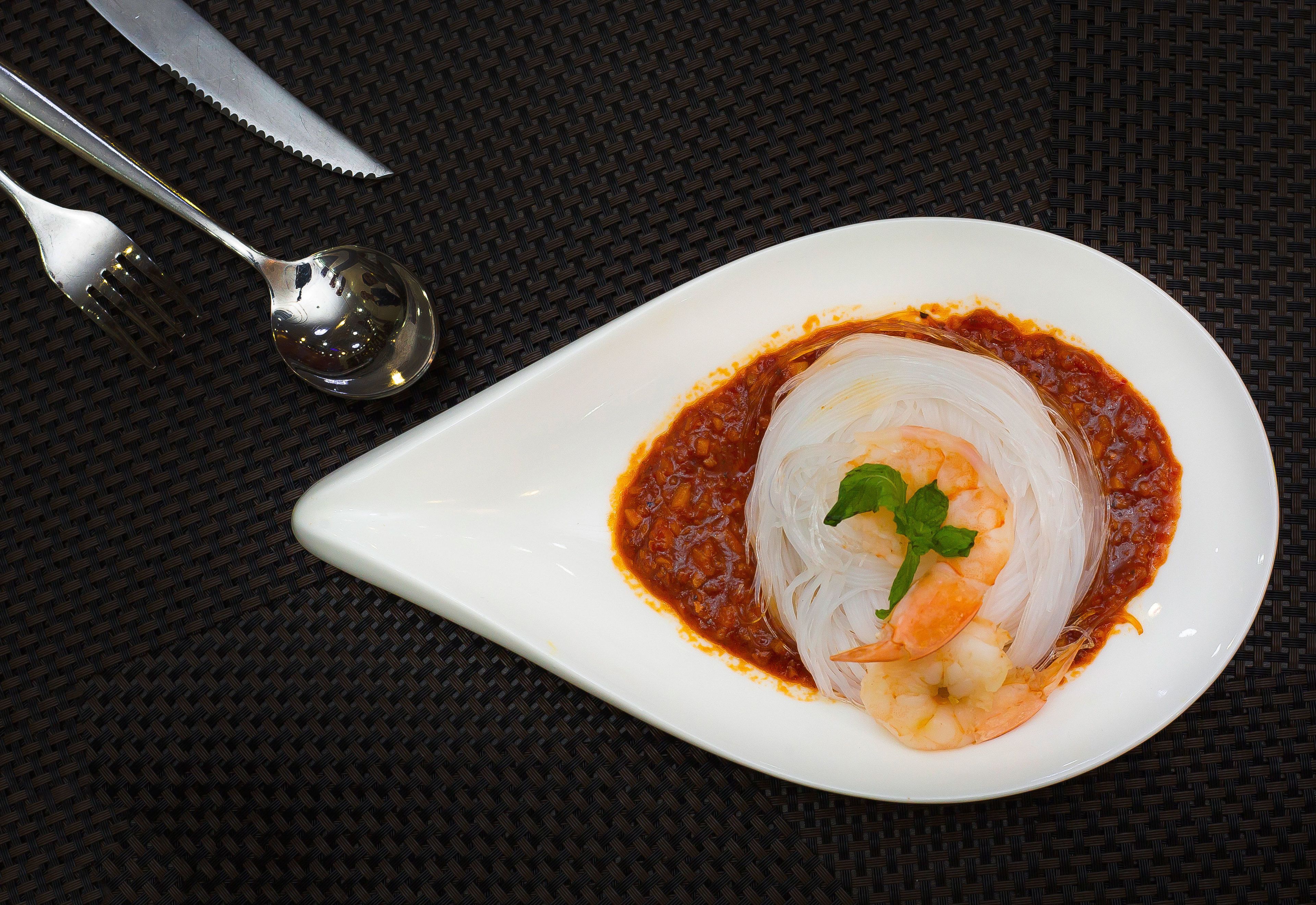 A white plate with a dish of noodles, shrimp, and sauce, accompanied by a fork and spoon on a dark woven surface.