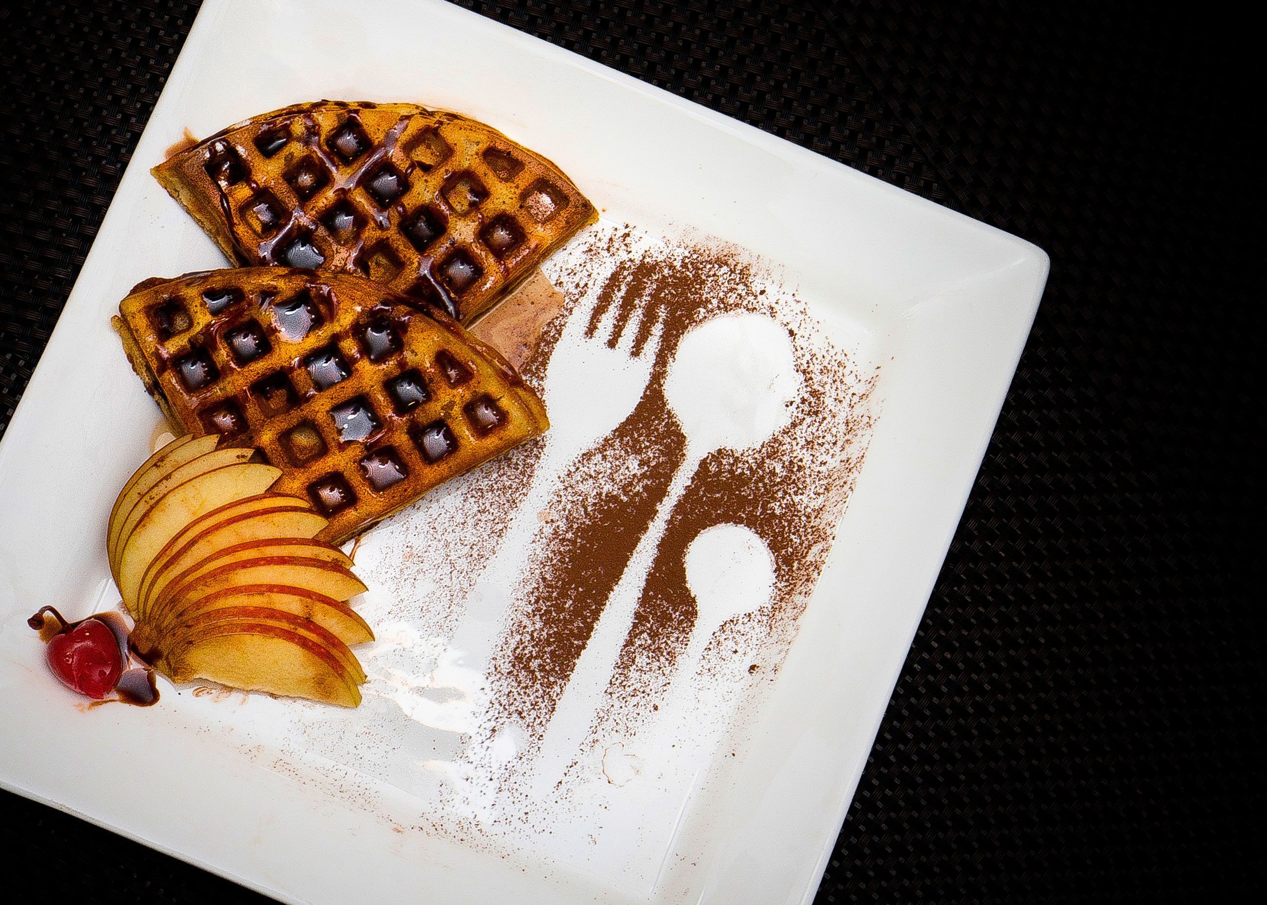 A square plate with three waffles, apple slices, and a dusting of cinnamon.