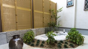 Un jardín con un jarrón decorativo, un árbol pequeño y una pared blanca con ventanas.