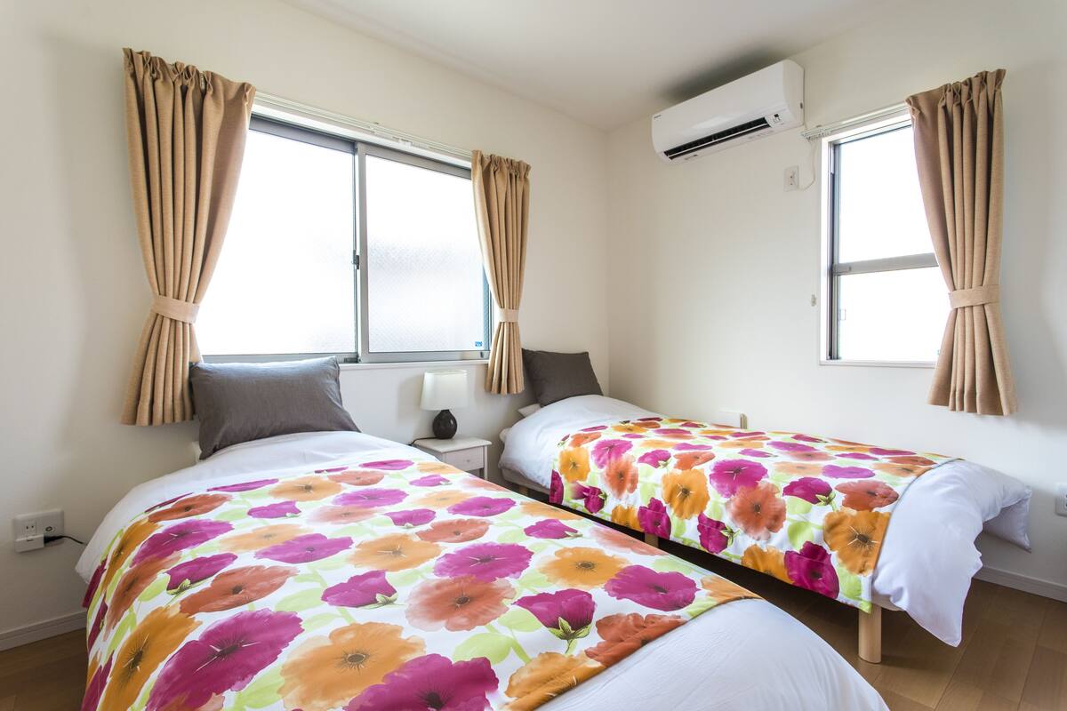 A room with two beds, each with floral bedding, a window with curtains, a bedside table with a lamp, and an air conditioning unit.