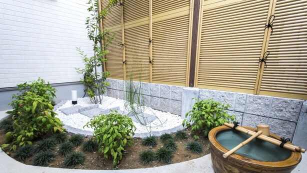 Un jardín japonés tradicional con una pila de piedra, valla de bambú y diversas plantas.
