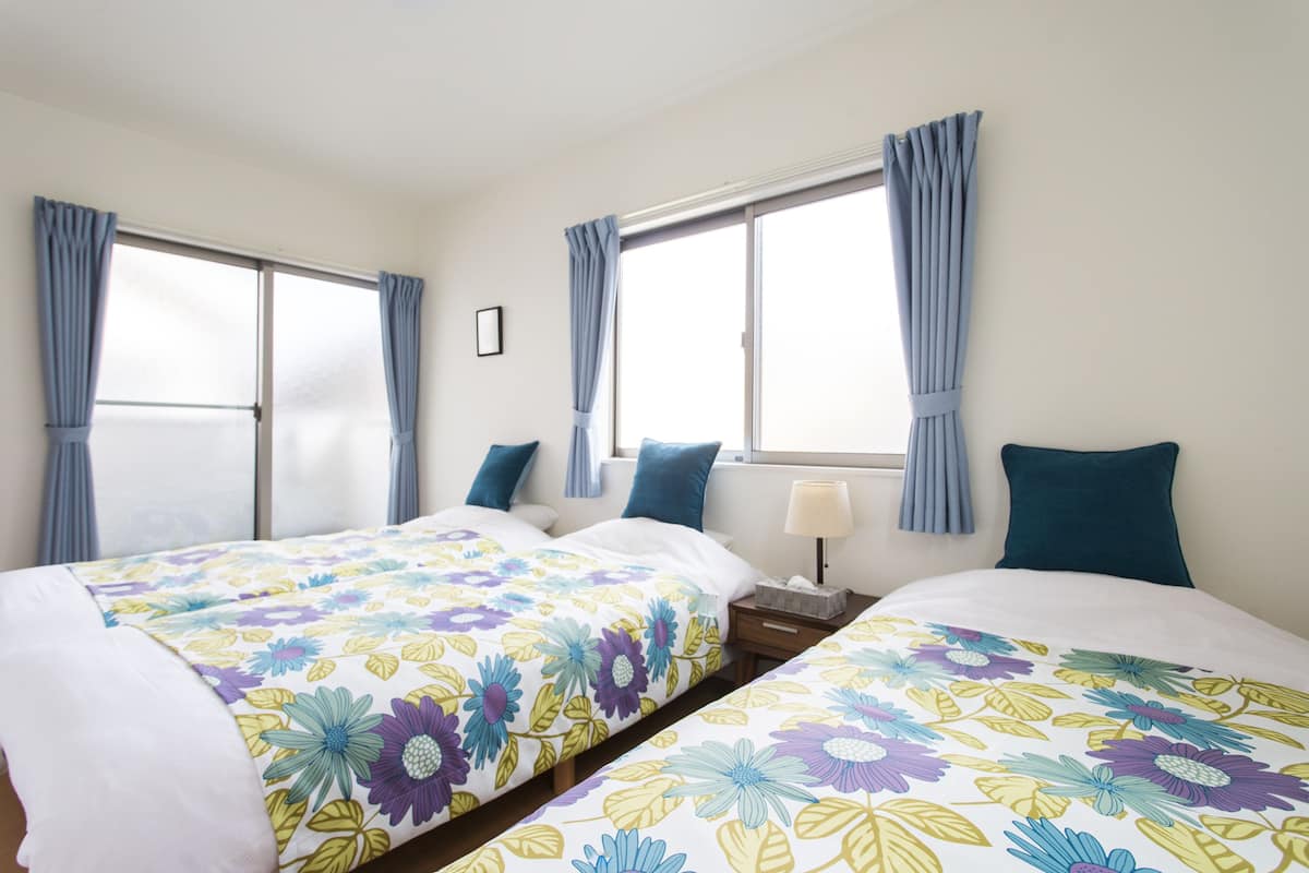 A hotel room with two beds, each with floral bedding, a nightstand with a lamp, and large windows with curtains.