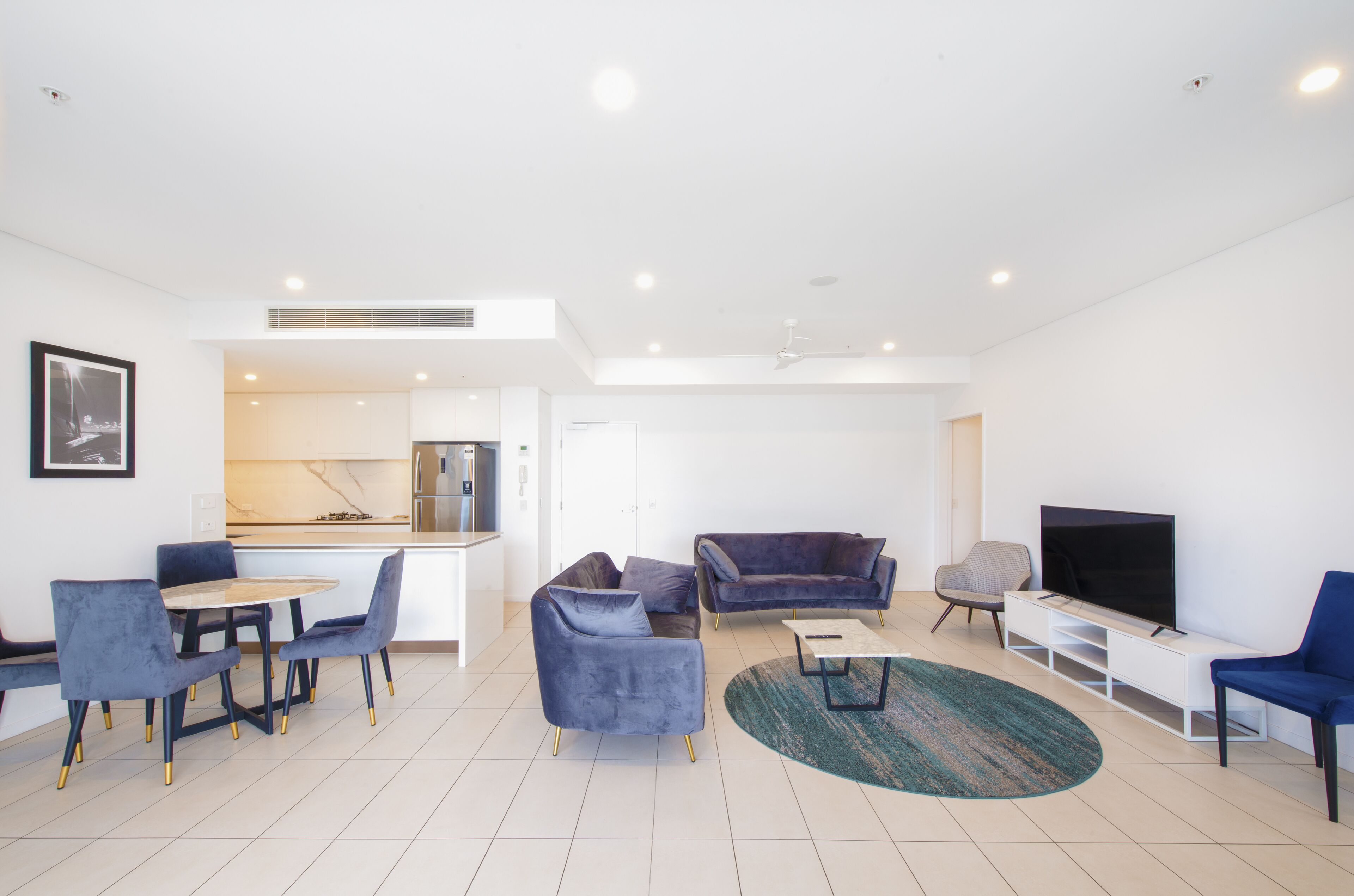 A modern living room with a dining area, a kitchen in the background, and a flat-screen TV.