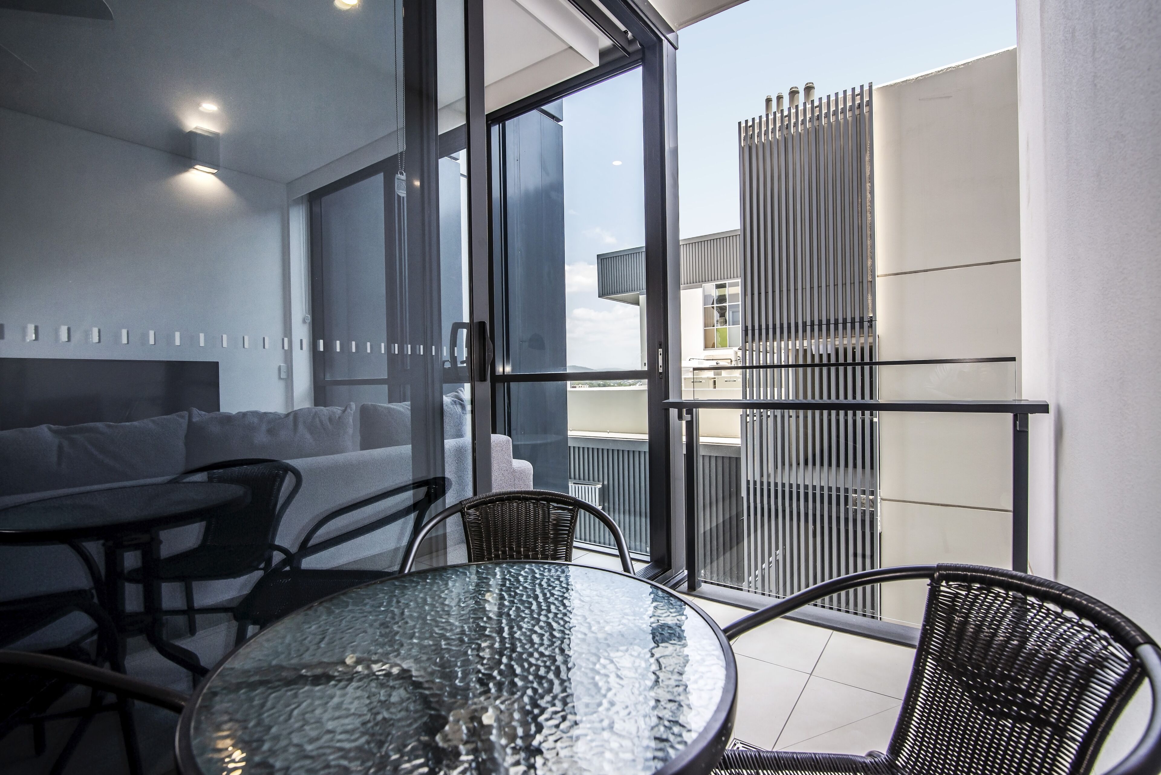 Un balcón moderno con puerta de cristal, una mesa redonda de cristal y sillas de mimbre negro.