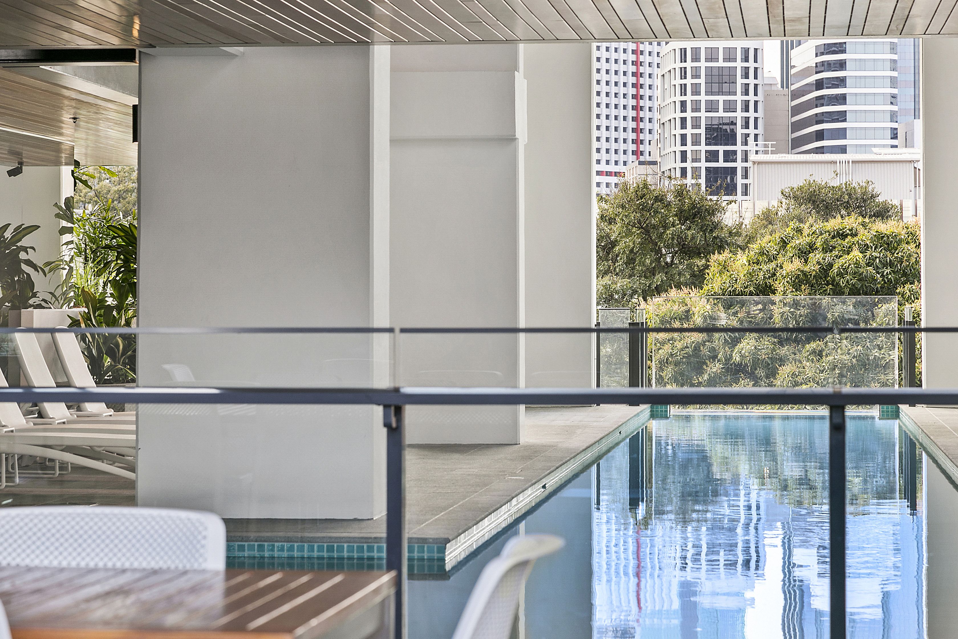A modern hotel balcony with a pool, lounge chairs, and a view of city buildings.