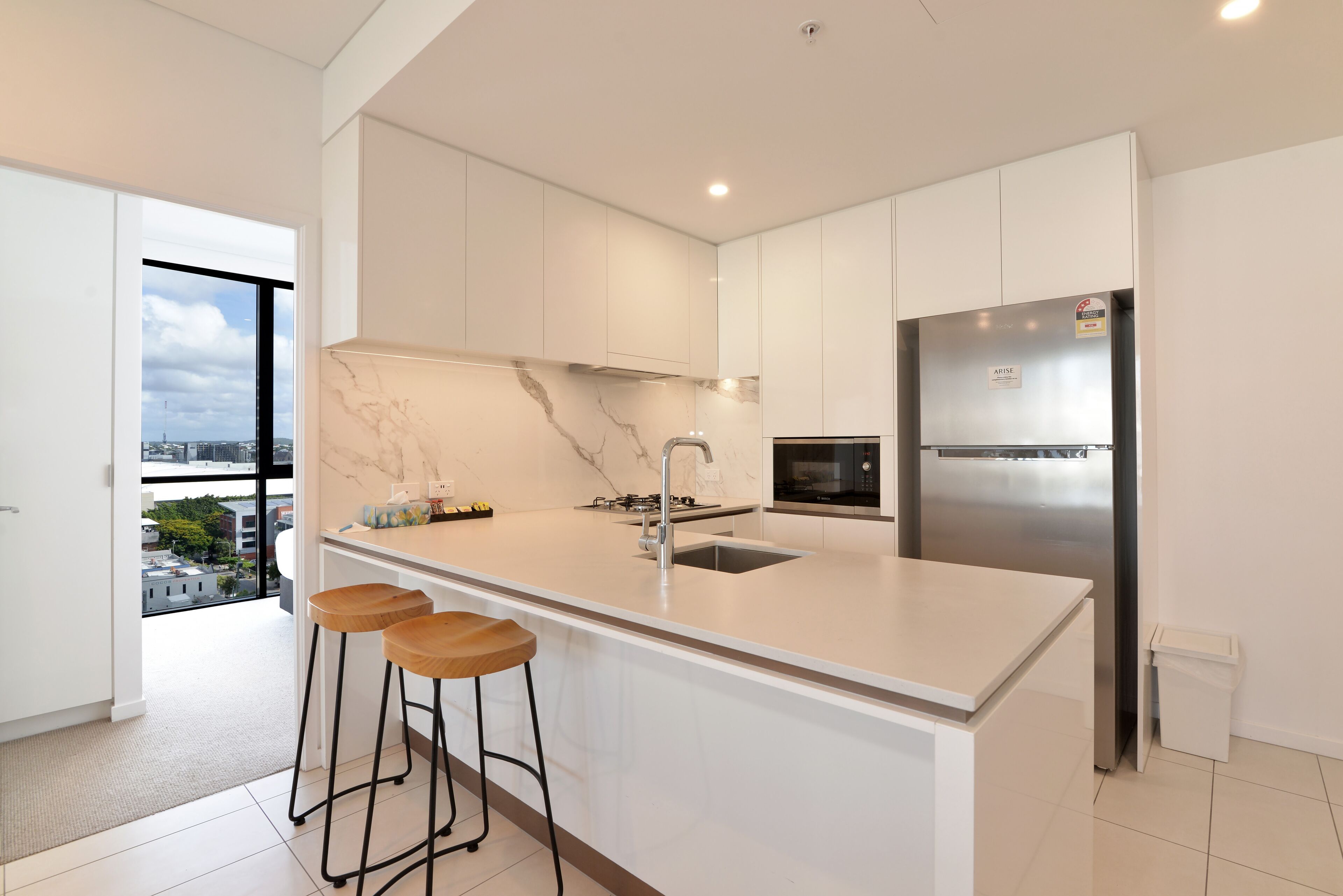 A modern kitchen with white cabinetry, a marble backsplash, a stainless steel refrigerator, and a large island with bar stools.