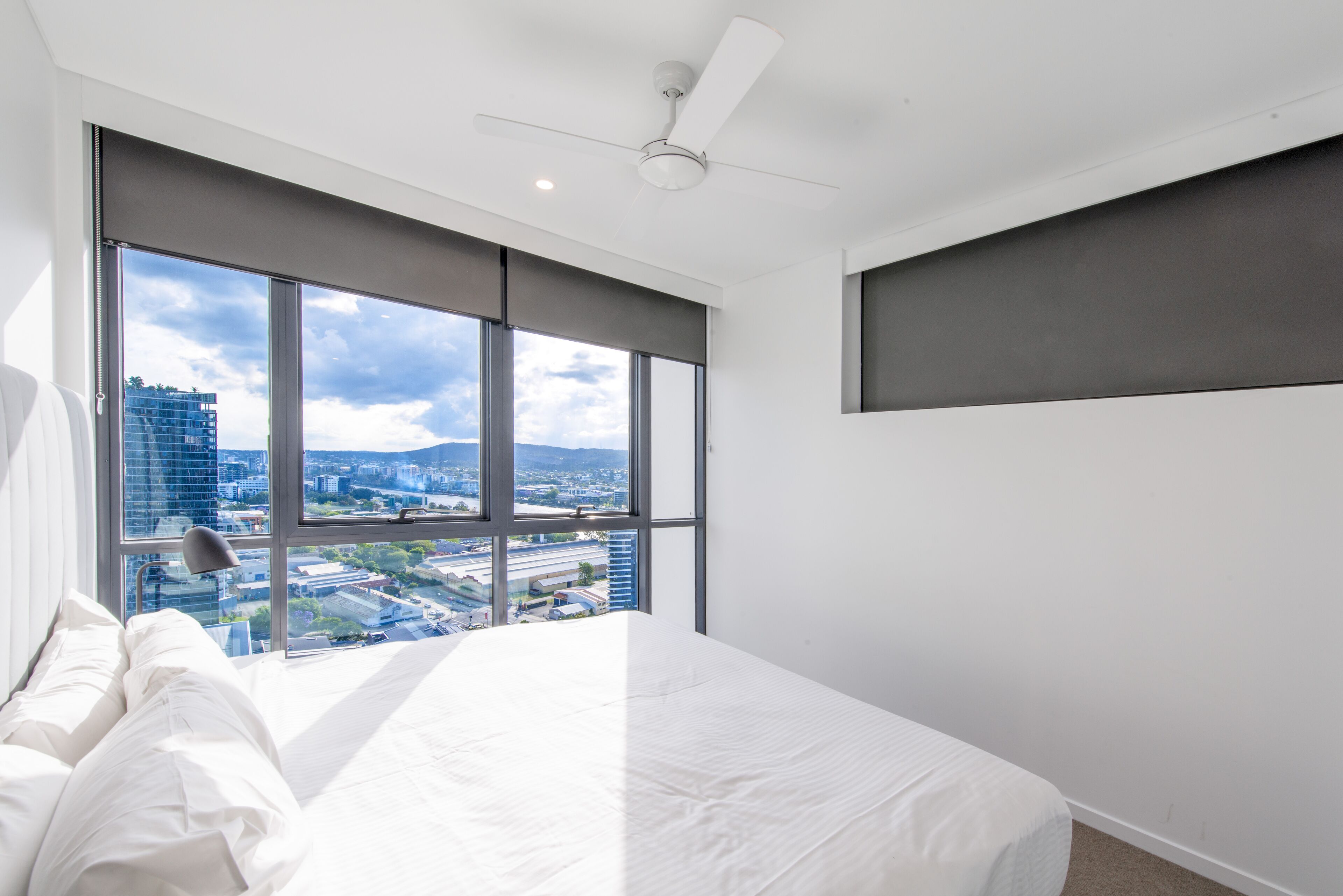 A modern hotel room with a large bed, a ceiling fan, and a city view through floor-to-ceiling windows.