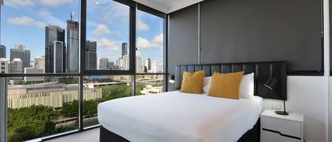 A modern hotel room with a large bed, a nightstand, and a view of a city skyline through floor-to-ceiling windows.